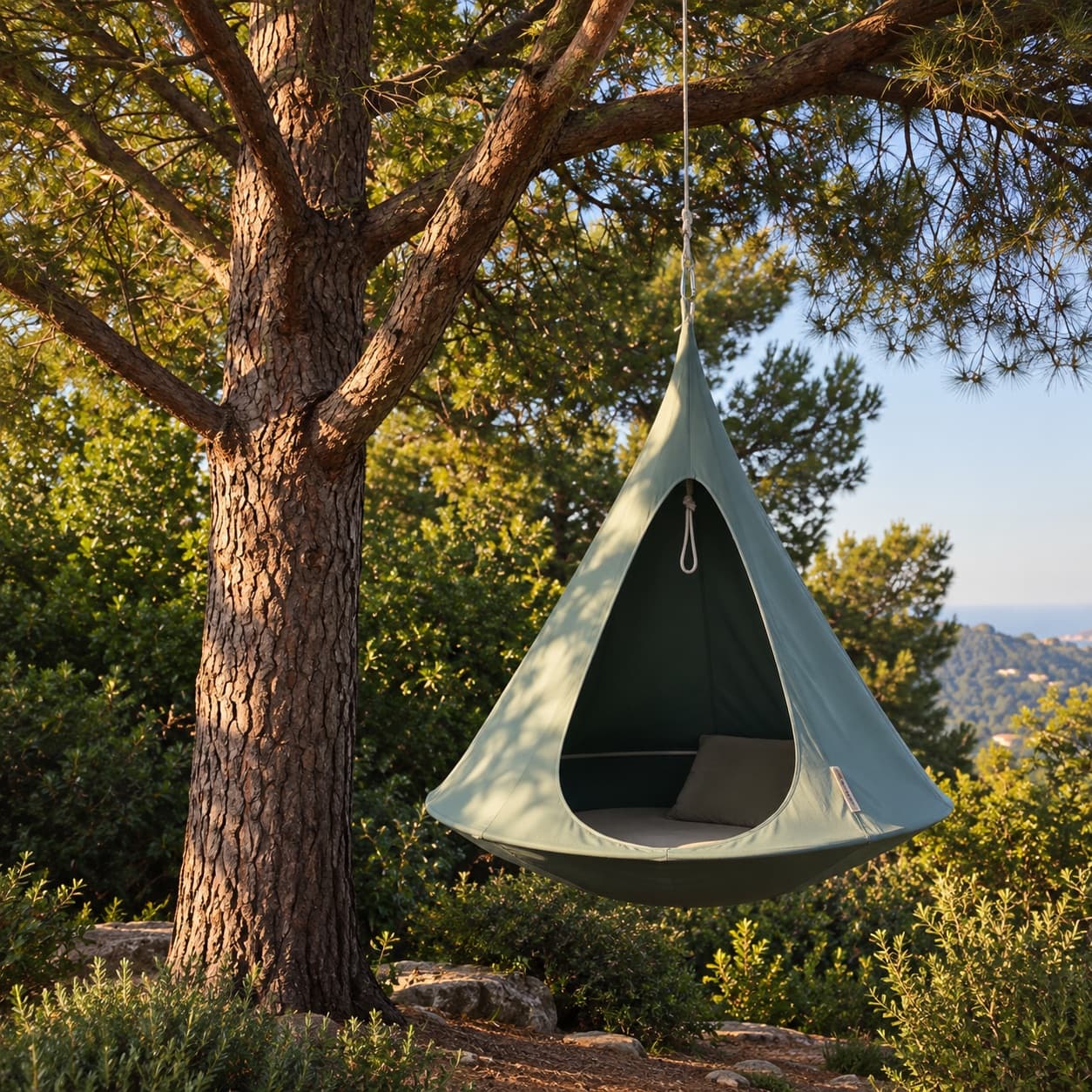 A green canvas tent hanging from a pine branch, with a cushion inside and the hills behind